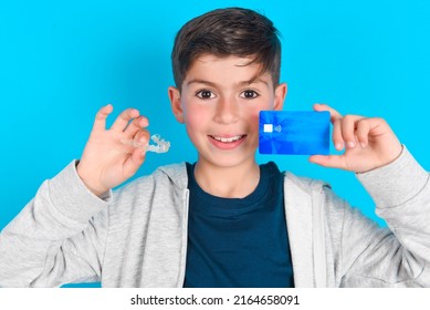 Brunette Kid Boy Wearing Grey Hoodie Over Blue Background Holding A Credit Card And Invisible Aligner.  Ready To Finance Treatment