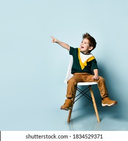 Brunette Kid In Colorful T-shirt And Brown Pants. Smiling And Pointing At Something By Forefinger, Sitting On White Chair Against Blue Studio Background. Childhood, Fashion. Close Up, Copy Space