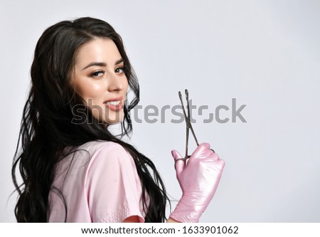 Brunette lady in pink suit and latex gloves posing standing sideways isolated on white. She is holding metal clamps. Medical staff, cosmetologist, hairdresser. Copy space, advertising area. Close up