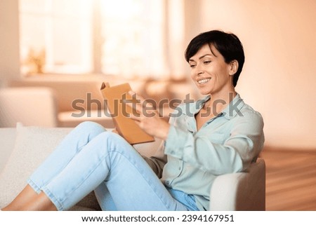 Brunette lady smiling absorbed in reading fiction, comfortably seated on her sofa with paper book, epitomizing peaceful weekend break, surrounded by the comfort of her home interior