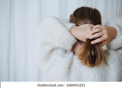 Brunette Little Girl In White Fluffy Knitted Sweater Putting Her Hair In A Pony Tail. At Home, In Front Of A Curtain. From Behind.