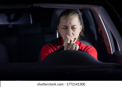 brunette long hair girl in red sweater driving car at night yawns tired holding steering wheel with one hand. safe driving concept. copy space. Lack of sleep fatigue concept