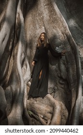 Brunette With Long Hair Posing In A 700 Years Old Tree. Women In Long Black Dress On Banyan Tree In Summer Day. Travel, Adventure, Eco Tourism, In Harmony With Nature. Bali, Indonesia.