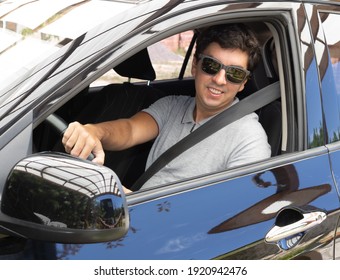 Brunette Man In A Black Car With A Seat Belt Wearing Sunglasses And A Gray Polo Shirt With A Collar. Driving In Holiday Mood And Good Vibes.