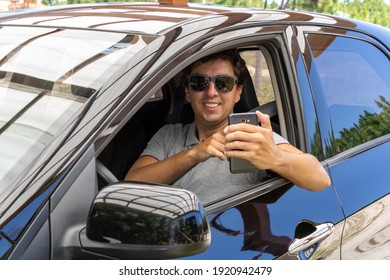 Brunette Man In A Black Car With A Seat Belt Wearing Sunglasses And A Gray Polo Shirt With A Collar. With A Mobile Phone Typing A Message To Friends. Driving In Holiday Mood And Good Vibes.