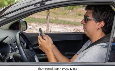 Brunette Man In A Black Car With A Seat Belt Wearing Sunglasses And A Gray Polo Shirt With A Collar. With A Mobile Phone Typing A Message To Friends. Driving In Holiday Mood And Good Vibes.