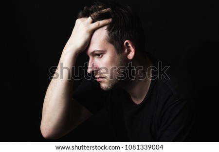A brunette man with a bristle in a black T-shirt on a dark background holds his head in one hand. View in profile. Social problems.