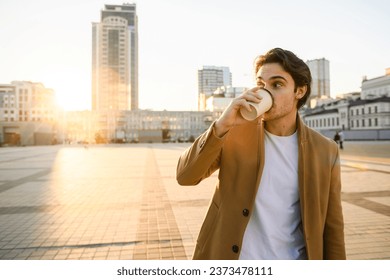 Brunette man in coat and white t-shirt drinking coffee against sunset in city	 - Powered by Shutterstock