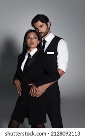 Brunette Man Looking At Camera While Embracing Asian Woman In Black Blazer Dress On Grey Background