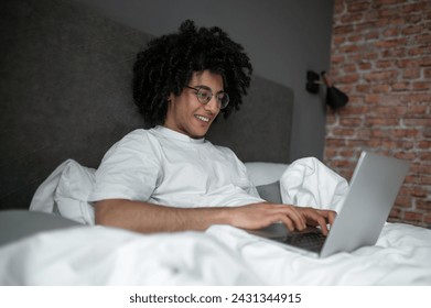 Brunette man in white tshirt lying in bed and working on laptop - Powered by Shutterstock