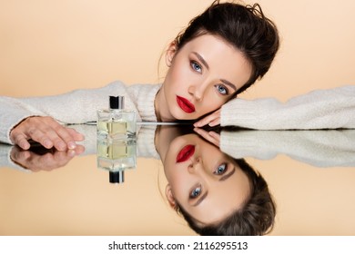 Brunette Model With Red Lips Looking At Camera Near Perfume On Mirror Isolated On Beige