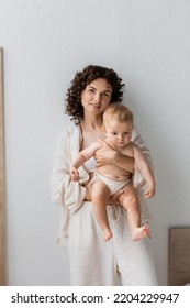 Brunette Mom In Pajamas Holding Baby Daughter And Looking At Camera At Home