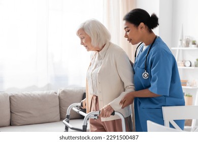 brunette multiracial nurse in uniform supporting retired woman with grey hair walking with walker at home - Powered by Shutterstock