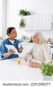 brunette multiracial nurse using digital tablet and holding medication while talking to retired woman with grey hair - Shutterstock ID 2291500377