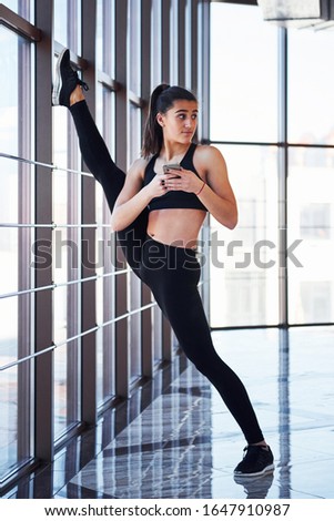 Brunette with phone in hands leaning on the railings of window and doing long stretching for legs.