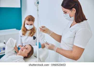 Brunette Physician In White Uniform And Gloves Looking To The Intravenous Vitamin Drip In Spa Salon