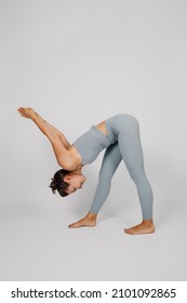 Brunette Poses On Isolated Background. Young Woman In Gray Yoga Tracksuit With Her Hair In Bun Is Doing Stretching Exercise, She Leaned Forward And Stretched Her Arms Up, Side View.