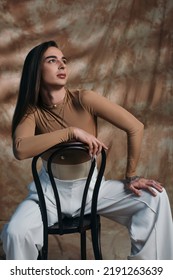 Brunette Queer Person In White Pants Sitting On Chair On Abstract Brown Background