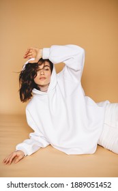 Brunette with short curly hair posing on beige background. She is dressed in white hoodie and jeans. Sexy woman touching face. Winter, fall or spring outfit. Sexy woman wear white total look. 