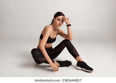 brunette sportswoman in black active wear with fitness tracker on wrist sitting on grey backdrop - Powered by Shutterstock