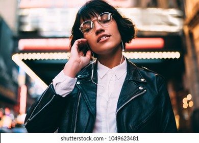 Brunette stylish hipster girl with close eyes enjoying listening electronic music in earphones feeling carefree on free time standing in night city downtown of New York with neon illumination lights - Powered by Shutterstock