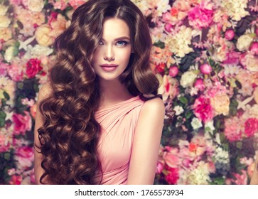 Brunette Summer Beauty Girl  Long  And   Shiny Wavy Hair .  Beautiful  Model Woman With Curly Hairstyle ,   Background  Wall Of Flowers .