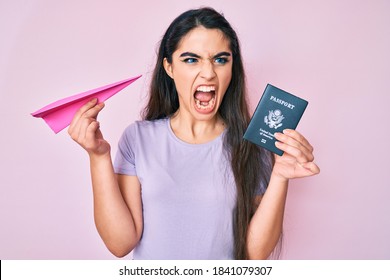 Brunette Teenager Girl Holding Paper Airplane And Usa Passport Angry And Mad Screaming Frustrated And Furious, Shouting With Anger. Rage And Aggressive Concept. 