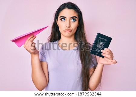 Brunette teenager girl holding paper airplane and usa passport smiling looking to the side and staring away thinking. 
