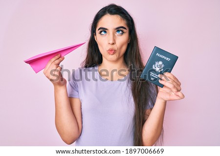 Brunette teenager girl holding paper airplane and usa passport making fish face with mouth and squinting eyes, crazy and comical. 