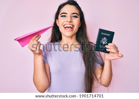 Brunette teenager girl holding paper airplane and usa passport smiling and laughing hard out loud because funny crazy joke. 