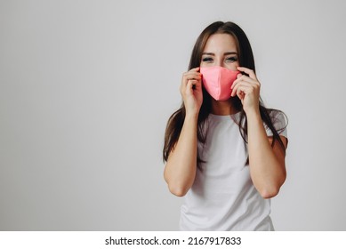 Brunette Wearing Pink Mask On White Background Smiling Happily. Girl Corrects Protective Mask On Her Face. Conceptual Administrator Wears Medical Mask
