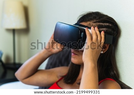 Brunette woman with braided hair positioning her virtual reality goggles to start the experience. Young girl in her room initiating an augmented reality visualization through her digital device.