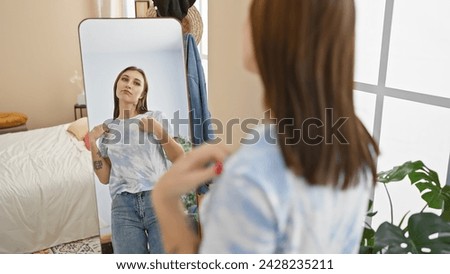 A brunette woman in casual attire adjusts her t-shirt in front of a mirror in a well-lit bedroom interior.
