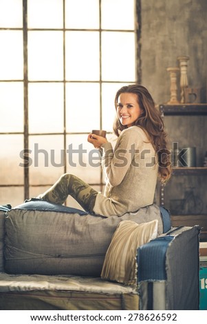 A brunette woman in comfortable clothing is is smiling and holding a hot cup of coffee, sitting on the back of a sofa. Industrial chic background, and cozy atmosphere. Loft decoration details.