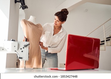 Brunette woman is considering a version of a dress on a mannequin and verifies with a pattern open on a laptop - Powered by Shutterstock