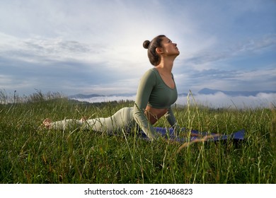 Brunette woman doing Surya Namaskar in mountains among green grass, looking at sky. Pretty female have practice in morning, standing in Bhujangasana, Cobra pose.