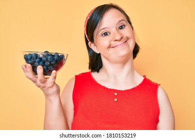 Brunette woman with down syndrome holding blueberries looking positive and happy standing and smiling with a confident smile showing teeth 