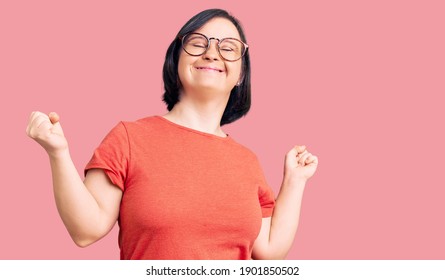 Brunette woman with down syndrome wearing casual clothes and glasses very happy and excited doing winner gesture with arms raised, smiling and screaming for success. celebration concept. 