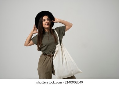 Brunette woman in green t-shirt and black hat holding cotton shopper bag with vegetables, products in white room. Eco friendly shopping bags. Zero waste, plastic free concept.