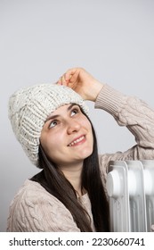 A brunette woman in a hat hugs an oil heater, warms up in winter.