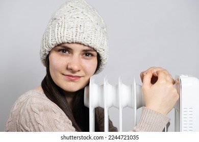 A brunette woman in a hat hugs an oil heater, warms up in winter.