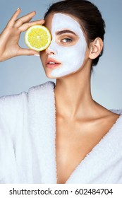 Brunette Woman Holding A Slice Of Lemon In Front Of Her Face. Photo Of Woman With Moisturizing Facial Mask. Beauty & Skin Care Concept