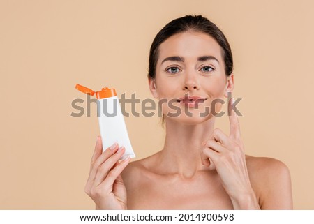 brunette woman holding sunscreen and pointing up with finger isolated on beige