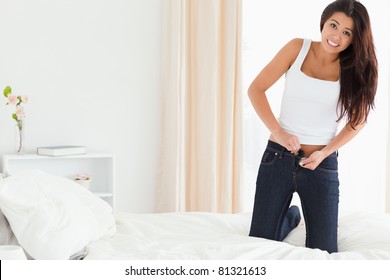 Brunette Woman Kneeing On Bed Trying To Close Her Jeans Looking Into Camera In Bedroom