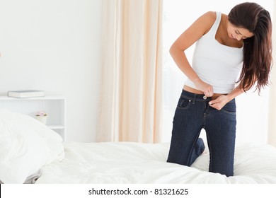 Brunette Woman Kneeing On Bed Trying To Close Her Jeans In Bedroom