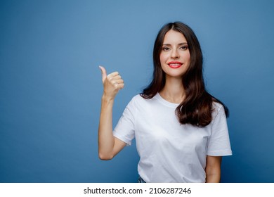 Brunette Woman With Long Hair In A White T-shirt Points Her Thumb To The Side. Female Portrait Isolated Against Blue Solid Background.