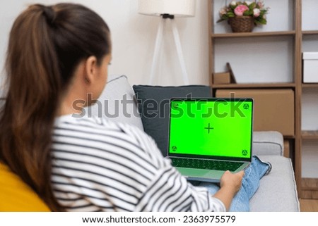 Brunette woman with long ponytail rests looking at laptop screen with chromakey female browses social media relaxing on sofa in light room at home backside view