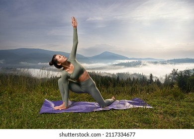 Brunette woman looking up doing Utthita Trikonasana on knees among mountains nature. Pretty relaxing woman have practice of yoga at sunrise time, deeply breathing. Concept of yoga.