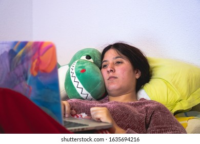 Brunette Woman Lying In Bed With A Computer And Holding A Stuffed T Rex Dinosaur In A Bedroom
