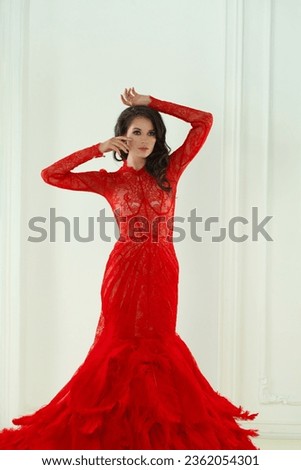 Brunette woman with makeup and long curly hairstyle wearing red evening gown in white interior. Vogue style model